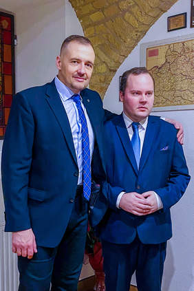 Pokaż zdjęcie: dwaj mężczyźni stoją obok siebie ubrani w granatowe garnitury, starszy położył swoją dłoń na ramieniu młodszego.