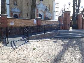 Pokaż zdjęcie: Przy murze ogrodzeniowym z cegły klinkierowej koloru miodowego jest na całej długości podjazd. Poręcze są w kolorze czarnym. W tle jest budynek kościoła.