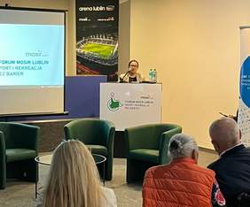 Pokaż zdjęcie: Konferencja, przy mównicy stoi kobieta w okularach. 