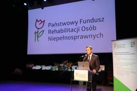 Pokaż zdjęcie: Ubrany w garnitur mężczyzna, na tle logo PFRON i baneru CIDON odczytuje list gratulacyjny