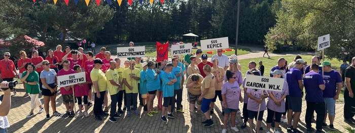 Pokaż zdjęcie: Słoneczna pogoda. W rzędzie stoi kilkadziesiąt osób niepełnosprawnych, pogrupowanych w kolorów koszulek. W każdej grupie ktoś trzyma tablicę z nazwą swojej placówki.