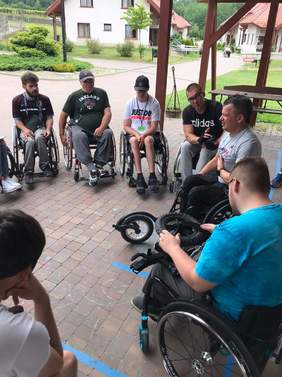 Pokaż zdjęcie: grupa uczestników obozu poruszająca się na wózkach uczestniczy w zajęciach na otwartej przestrzeni  
