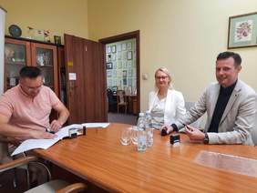 Pokaż zdjęcie: Przy owalnym stole siedzą trzy osoby: kobieta i dwóch mężczyzn. Jeden z panów przystawia pieczątkę do dokumentów. W tle widać ścianę, na której wisi bardzo dużo dyplomów i gratulacji. 