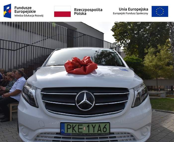 Pokaż zdjęcie: Na górze, od lewej strony zamieszczone zostały następujące logotypy: Funduszy Europejskich – Programu Wiedza-Edukacja-Rozwój; Rzeczpospolitej Polskiej oraz Unii Europejskiej – Europejskiego Funduszu Społecznego. Poniżej widoczny jest biały bus z czerwoną kokardą na masce i naklejką informującą, że pojazd przystosowany jest do przewozu osób z niepełnosprawnością. W tle można dostrzec drzewa liściaste, schody prowadzące do budynku o płaskim dachu oraz siedzące na krzesłach osoby, które biorą udział w uroczystym przekazaniu mikrobusa.