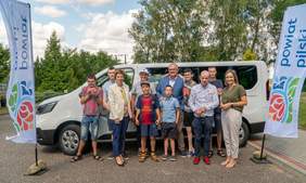 Pokaż zdjęcie: Grupa jedenastu osób, zarówno dorosłych, jak i dzieci, stoi na parkingu, na tle białego busa z naklejką z logo Państwowego Funduszu Rehabilitacji Osób Niepełnosprawnych informującą, że pojazd dofinansowany jest ze środków PFRON. Po obu stronach busa widoczne są dwie duże chorągiewki z logotypem powiatu pilskiego. W oddali możemy dostrzec duże skupisko drzew liściastych oraz iglastych.