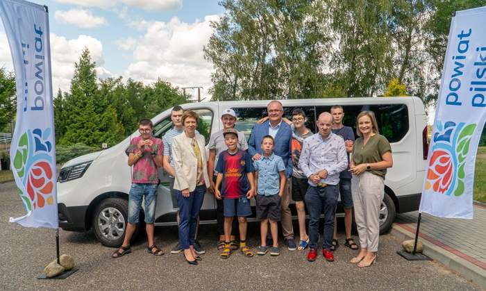 Pokaż zdjęcie: Grupa jedenastu osób, zarówno dorosłych, jak i dzieci, stoi na parkingu, na tle białego busa z naklejką z logo Państwowego Funduszu Rehabilitacji Osób Niepełnosprawnych informującą, że pojazd dofinansowany jest ze środków PFRON. Po obu stronach busa widoczne są dwie duże chorągiewki z logotypem powiatu pilskiego. W oddali możemy dostrzec duże skupisko drzew liściastych oraz iglastych.