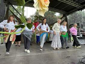 Pokaż zdjęcie: Kobiety w różnym wieku ubrane w barwne stroje tańczą na dużej scenie udekorowanej sztucznymi kwiatami o wielkich rozmiarach. Z tyłu sceny zawieszone są balony w kształcie liczby „25”. W tle wysokie drzewa, a pośród nich kwitnące kasztany.