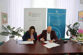 Pokaż zdjęcie: Uroczyste podpisanie umowy, w gabinecie za stołem konferencyjnym kobieta i mężczyzna podpisują umowę.