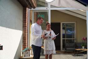 Pokaż zdjęcie: dwie osoby od lewej mężczyzna w białej koszuli, obok kobieta w białej sukience, trzymają mikrofony,uśmiechają się