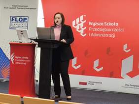 Pokaż zdjęcie: Przy mównicy stoi kobieta ubrana w czarny garnitur, w tle dużo koloru czerwonego z logo Wyższej Szkoły Przedsiębiorczości i Administracji w Lublinie.