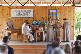 Pokaż zdjęcie: Na scenie występ zespołu śpiewaczego, zespół przebrany w długie suknie w kolorze brązowym, kobiety mają wianki na głowach.