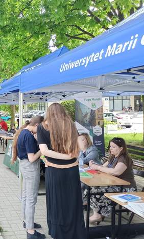 Pokaż zdjęcie: Dwie kobiety stoją tyłem przy stoisku na pikniku informacyjnym, oglądają materiały promocyjne