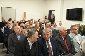 Pokaż zdjęcie: Zdjęcie osób siedzących na krzesełkach w sali konferencyjnej nowego budynku.