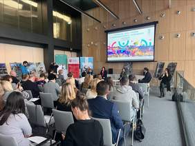 Pokaż zdjęcie: Zdjęcie sali konferencyjnej robione z tyłu sali, uczestnicy siedzą na krzesłach, na ścianie wyświetlana jest prezentacja.