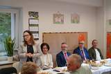 Spotkanie przy wspólnym stole w świetlicy. Kilka osób siedzi przy poczęstunku, a jedna osoba stoi i przemawia. Na ścianach widoczne są prace plastyczne dzieci.