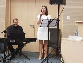 z lewej strony mężczyzna siedzi i gra na instrumencie klawiszowym, z prawej strony kobieta w białej sukience spiewaa