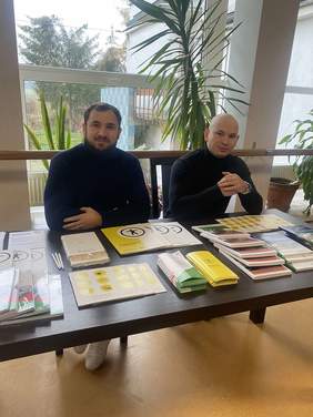 Pokaż zdjęcie: Dwóch mężczyzn siedzi przy stole z ulotkami, broszurami, informatorami. Znajdują się wewnątrz budynku, za nimi kilka kwiatów oraz duże okna. Na zewnątrz widać drzewa oraz balkon.