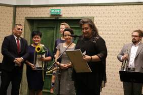 Pokaż zdjęcie: Na scenie stoi kobieta w ciemnych długich włosach zaczesanych do tyłu. Ubrana jest w ciemny kostium z wstawkami z ciemnej siateczki. Patrzy na trzymany oprawiony dyplom. Z tyłu stoją pozostali laureaci oraz osoby wręczające wyróżnienie.