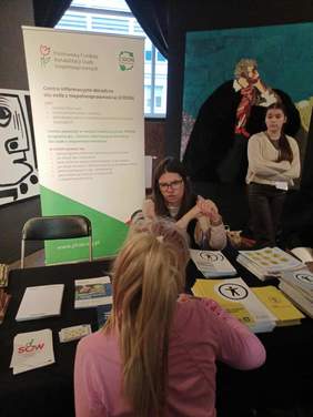 Dwie młode kobiety, siedzą przy stole, rozmawiają, w tle biało-zielony baner z napisem CIDON, na stole leżą ulotki