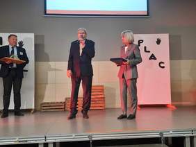 Pokaż zdjęcie: Na scenie stoją trzy osoby: jedna kobieta i dwóch mężczyzn. Pan pośrodku przemawia do mikrofonu. Wszyscy są elegancko ubrani. 