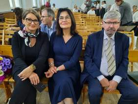 Pokaż zdjęcie: W pierwszym rzędzie siędzą dwie kobiety i jeden mężczyzna. Uśmiechają się szeroko. Za nimi widać inne osoby wchodzące do sali gimnastycznej. 