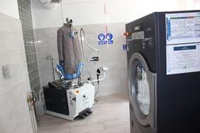 Pokaż zdjęcie: W wykafelkowanym pomieszczeniu po prawej stronie stoi bardzo duża pralka, w lewym rogu stoi urządzenie do prasowania na które nałożony jest płaszcz.