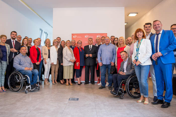 Pokaż zdjęcie: Uczestnicy uroczystości stoją wszyscy obok siebie na tle ścianki Krajowej Izby Fizjoterapuetów w koloerze czerwonym, w małym pomieszczeniu z biayłmi ścianami, są też osoby na wózkach inwalidzkich