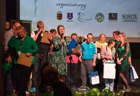 Pokaż zdjęcie: Na scenie stoją artyści oraz organizatorzy. Artyści trzymają w rekąch upominki, m.in. od PFRON