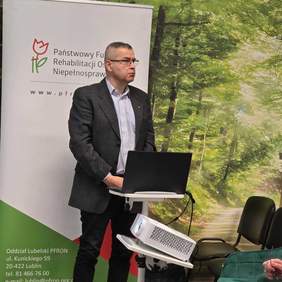Pokaż zdjęcie: Przy laptopie stoi mężczyzna w garniturze, w tle rollup z logo PFRON.