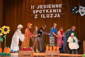 Pokaż zdjęcie: Na scenie dzieci odgrywają scenkę z Rzepki, ustawiły się jedno za drugim i ciągną pierwszą osobę siedzącą na krześle.