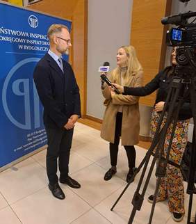 Inspektor PIP-y Grzegorz Meler podczas udzielania wywiadu, za nim stou baner Inspekcji, a obok niego, na prawo, redaktorki z mikrofonami