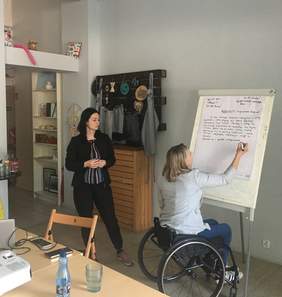Pokaż zdjęcie: Kobieta na wózku pisze markerem na flipcharcie stojącym w jasnym pomieszczeniu. Druga kobieta stoi obok i obserwuje.