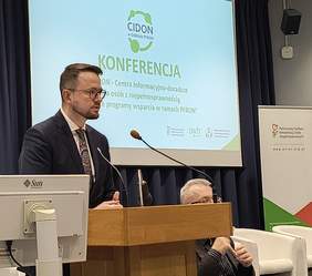 Pokaż zdjęcie: Dyrektor w okularach, ubrany w granatowy garnitur przy mównicy w auli politechniki