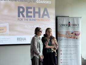 Pokaż zdjęcie: Dwie kobiety stoją przed ekranem z prezentacją konferencji REHA FOR THE BLIND IN POLAND. Jedna z nich mówi do mikrofonu. Obok znajduje się roll-up Fundacji Szansa – Jesteśmy Razem.