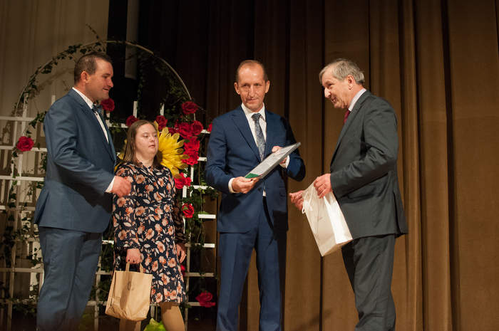 Pokaż zdjęcie: Na scenie stoi trzech mężczyzn w garniturach i kobieta w sukience w ręce trzyma papierową torebkę. Jeden z mężczyzn trzyma w rękach białą torebkę, a drugi trzyma w rękach list gratulacyjny w srebrnej ramce.