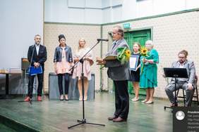 Pokaż zdjęcie: Mężczyzna stoi przy mikrofonie, za nim inni laureaci i orkiestra