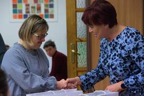 Pokaż zdjęcie: Dwie kobiety stoją na przeciw siebie trzymają się za dłonie , starsza kobieta składa gratulacje młodszej kobiecie.