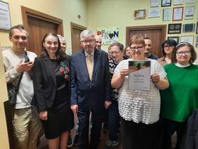 Pokaż zdjęcie: W siedzibie Łódzkiego Oddziału PFRON stoją ludzie. Wśród nich jest dyrektor Łódzkiego Oddziału PFRON oraz laureatka konkursu plasycznego. Zwyciężczyni prezentuje pamiątkowy dyplom, który otrzymała. 