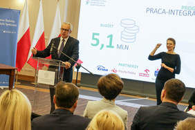 Pokaż zdjęcie: Mężczyzna w garniturze mówi do mikrofonu. W tle wyświetla sie prezentacja. Z prawej strony kobieta tłumacząca na język migowy.