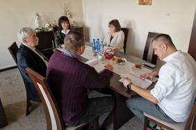 Pokaż zdjęcie: na zdjęciu duże pomieszczenie na środku stoi duży stół przy którym siedzą cztery osoby rozmawiają ze sobą na stole leży dokumentacja która jest omawiana przez osby zgromadzone w pomieszczeniu