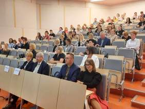 Pokaż zdjęcie: Na auli uniwersyteckiej siedzą w rzędach ludzie, którzy słuchają wykładu. Niektóre krzesła są wolne. 