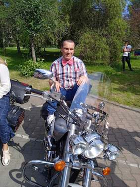 Pokaż zdjęcie: Na motorze siedzi młody mężczyzna ubrany w koszule w kratkę i czerwoną muchę pod szyją.