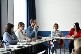 Pokaż zdjęcie: Siedzący przy stoliku mężczyzna mówi do mikrofonu. Siedzące obok osoby z uwagą słuchaja prelegenta.