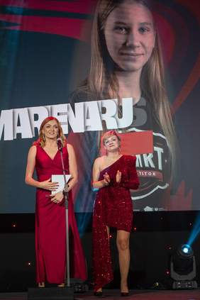 Pokaż zdjęcie: Dwie kobiety stoją na scenie podczas gali sportowej. Jedna z nich, ubrana w długą czerwoną suknię, stoi przy mikrofonie, druga w krótkiej czerwonej, błyszczącej kreacji – klaszcze. W tle na dużym ekranie wyświetlone jest zdjęcie młodej sportsmenki oraz grafika z jej imieniem i nazwiskiem.