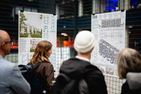 Pokaż zdjęcie: Grupa osób stojących tyłem przed dużymi tablicami informacyjnymi z rysunkami technicznymi i planem architektonicznym. W tle widoczna jest ściana z ciemnych płytek.