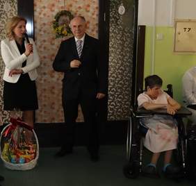 Pokaż zdjęcie: Trzy osoby, kobieta blondynka w czarnej sukience i białej marynarce trzyma w reku mikrofon, obok stoi mężczyzna w garniturze, dalej po prawej stronie siedzi kobieta na wózku inwalidzkim.