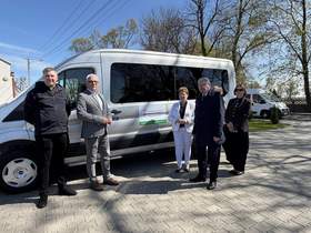 Przed samochodem stoi kilka osób. Wszyscy są elegancko ubrani. Na pojeździe jest naklejka z logo PFRON. 