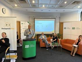 Pokaż zdjęcie: Mężczyzna stoi za mównicą i trzyma mikrofon w ręku. Po jego prawej stronie prezentacja wyświetlana na ekranie z logo PFRON. Obok dwie sofy dwuosobowe na których siedzą dwie kobiety i mężczyzna. Przed nimi stolik szklany z napojami. 