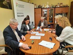 Pokaż zdjęcie: Przy owalnym stole siedzi pięć osób: trzy kobiety i dwóch mężczyzn. Są pochyleni nad umowami, które czytają i podpisują. Na stole leżą dokumenty, pieczątki, telefony, długopisy. 