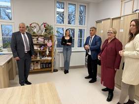 Pokaż zdjęcie: Pięć osób w pomieszczeniu z szafkami i regałem pełnym rękodzieła; mężczyźni w garniturach, kobiety w eleganckich strojach stoją i rozmawiają, za nimi duże okna z widokiem na drzewa.